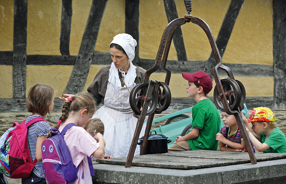 Frau in einem altertümlichen Gewand erklärt Kindern das alte Handwerk