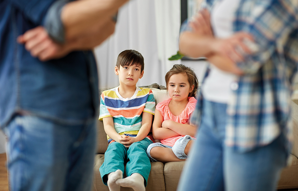 Ein Junge und ein Mädchen sitzen auf einem Sofa und schauen traurig auf ihre streitenden Eltern