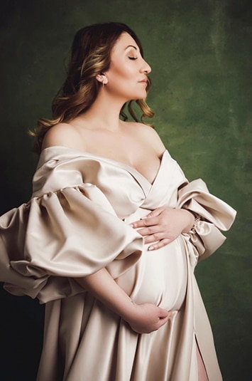 Eine schwangere Frau in einem cremefarbenen Kleid steht mit geschlossenen Augen und den Händen am Bauch seitlich zur Kamera